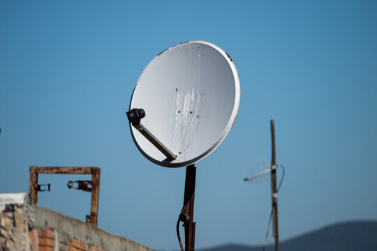 a weathered white satellite dish mounted on a rusty pole on a rooftop against a clear blue sky, with a blurred television antenna visible in the background.