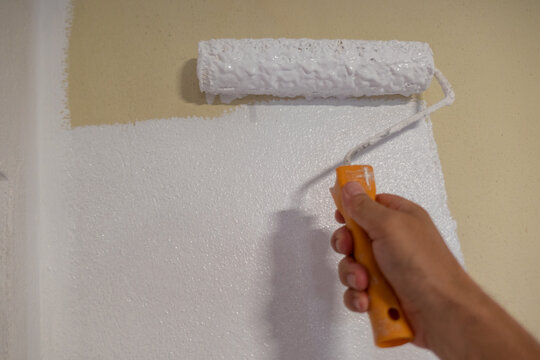 Pintor de rolo na m&atilde;o a aplicar tinta branca sobre a parede de cor creme durante a renova&ccedil;&atilde;o de interior de uma habita&ccedil;&atilde;o
