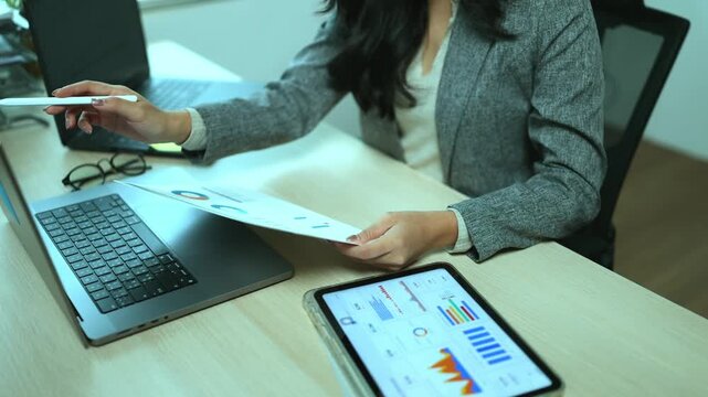 A professional Asian businesswoman wearing glasses is working intently at her office desk reviewing financial documents and data charts displayed on her laptop and tablet.
