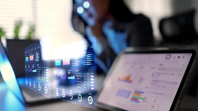 A focused Asian businesswoman analyzing financial data and business growth charts on a laptop and tablet with futuristic hologram analytics in a modern office.