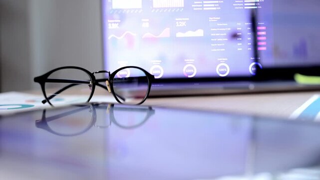 A modern workspace with a laptop displaying business analytics charts, a tablet, and a pair of glasses resting on a wooden desk, symbolizing financial analysis and corporate strategy.