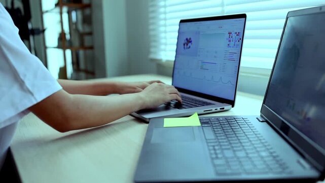 A female doctor in a white coat sitting at her desk, working and analyzing medical data, chemical structures, and graphs on dual laptops in a modern office or clinic setting.