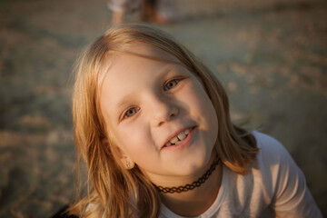 Fototapeta premium A young caucasian blonde girl smiling outdoors at sunset on sandy beach. Child looking at camera. Summer vacation. Childhood joy, happy kid, golden hour. High quality photo.