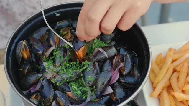 Close-up of a hand using a fork to pick mussels from a pot, with fries alongside, Cote d'Azur, France