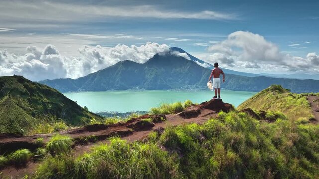Man take off white shirt standing on mountain peak, looking at expansive green tropical landscape of lake Batur and mount Agung. Active sport, achievement and travel in Bali. Aerial drone flight