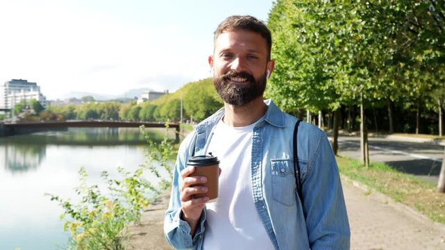 Young handsome man with a beard holding a cup of takeaway coffee and smiling at the camera while walking along a riverside path in a city park on a beautiful sunny day in slow motion