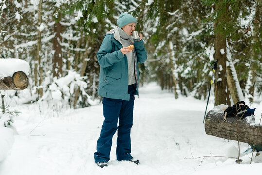 Forty-something lady having a winter snack break with a steaming drink and pastry in snowy forest. Mature woman by a log with a hot cup and sweet treat. Peaceful outdoor pause on a cold winter walk