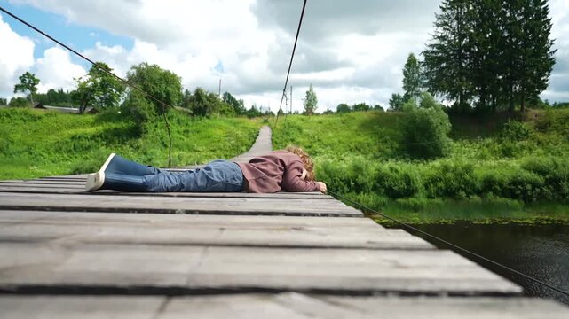 Carefree toddler rests on wooden bridge, gazing down at flowing river while enjoying rural landscape. This little boy pauses his journey to embrace serene countryside and peaceful outdoor atmosphere