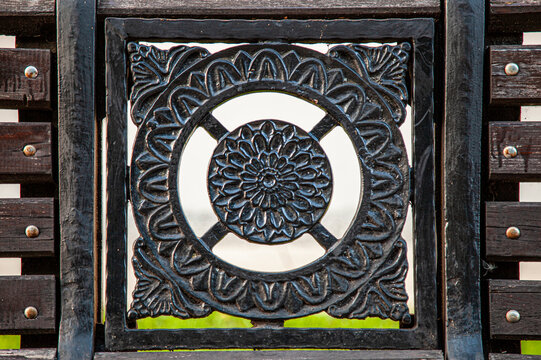 Close-up view of an ornate dark wrought iron architectural detail featuring floral scrollwork, geometric patterns, vertical bars, metal rivets, and weathered textures. 