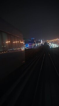 Oncoming metro train approaches and passes beside a forward moving train on an elevated railway, with city lights along the track at night