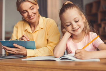 Obraz premium Smiling Caucasian Woman Helping Young Girl With Homework, Engaged In Learning Activity Together At Home. Joyful Interaction Between Female Adult And Child In Comfortable Indoor Setting.