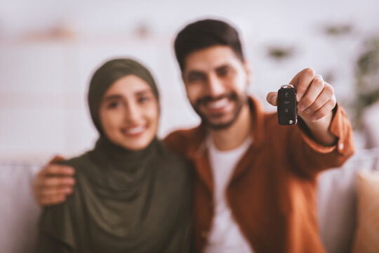 Joyful Young Couple, Smiling And Embracing Each Other While Showcasing Car Key At Home. They Are Excited About Their New Purchase, Sitting Comfortably In Their Living Room. Lifestyle Concept.