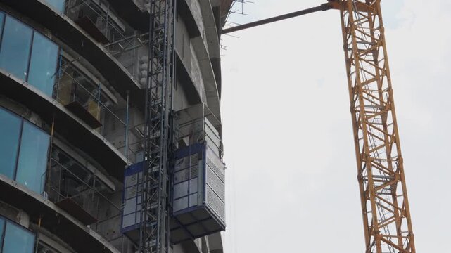Building hoist descends along an unfinished high rise beside a tower crane and suspended work platform