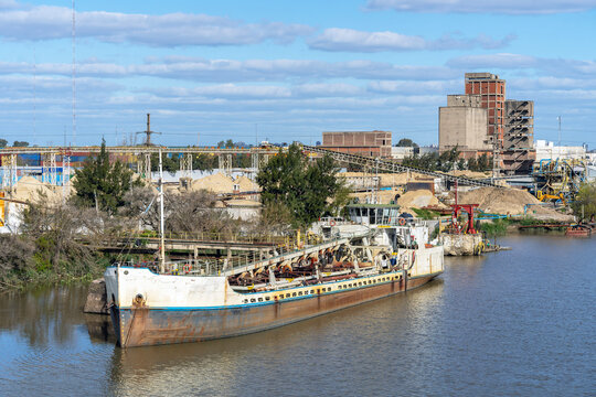 Industrial dredging vessel moored near riverbank with sand processing facility, conveyor systems, concrete factory, and industrial harbor infrastructure