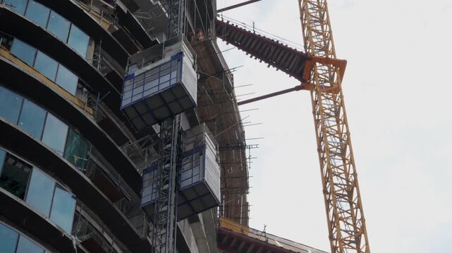 Construction hoist descends beside a tower crane on an unfinished high rise, revealing curved concrete floors, glass facade and scaffolding