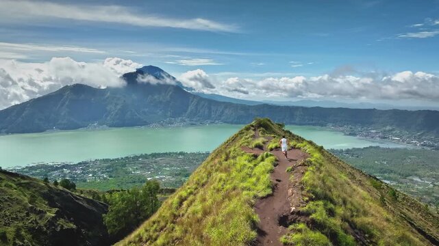 Man running on a grassy mountain ridge trail with mount Batur volcano and lake Batur in the background under a blue sky with clouds, showcasing adventure and travel in Indonesia. Aerial drone flight