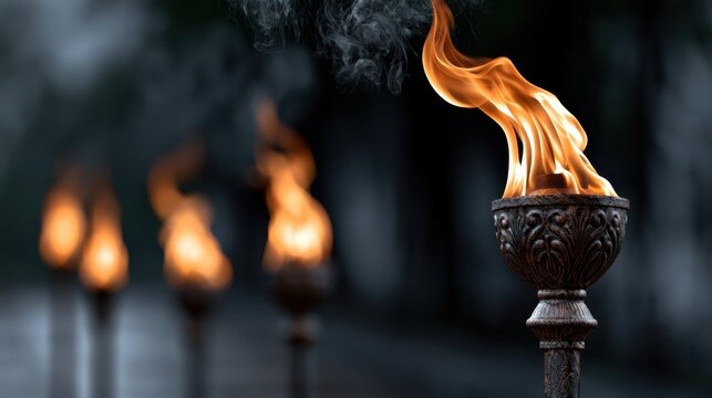 Ornate metal torch with bright orange flames glowing vividly in the nighttime with multiple torches blurred in the background for an atmospheric effect