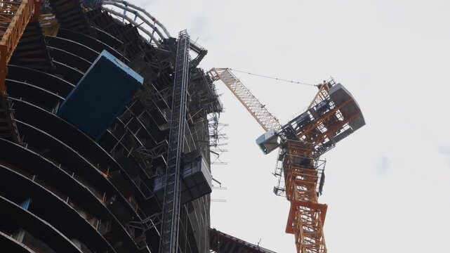 Tower crane stands beside an unfinished curved high rise, revealing concrete floors, scaffolding and a blue construction hoist