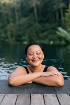 Smiling woman in black swimsuit relaxing at pool edge by a forest lake with eyes closed for wellness spa background and lifestyle banner