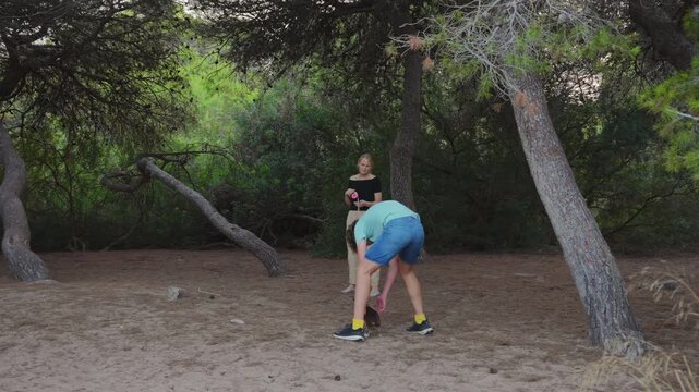 Mom and teenage son walk a small dog through a forest, the teenager bends down to pet the dog before they continue