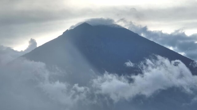 Mount Agung volcano peak emerges from thick blanket of morning flowing clouds, rays of light breaking through the overcast sky, highlighting the imposing natural landscape of Bali. Natural background