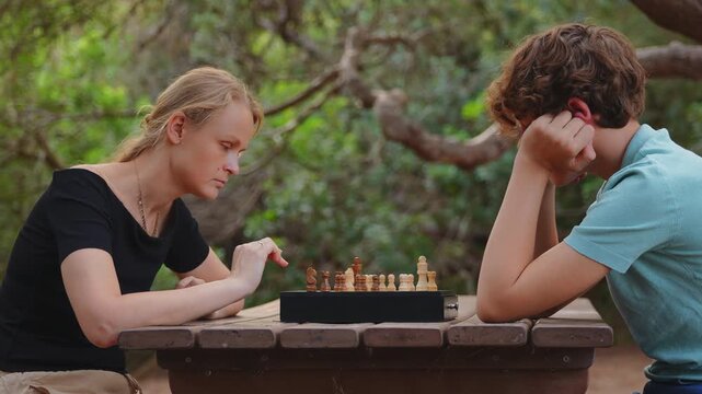 Mother makes a move in a chess game while her teenage son studies the board at an outdoor table