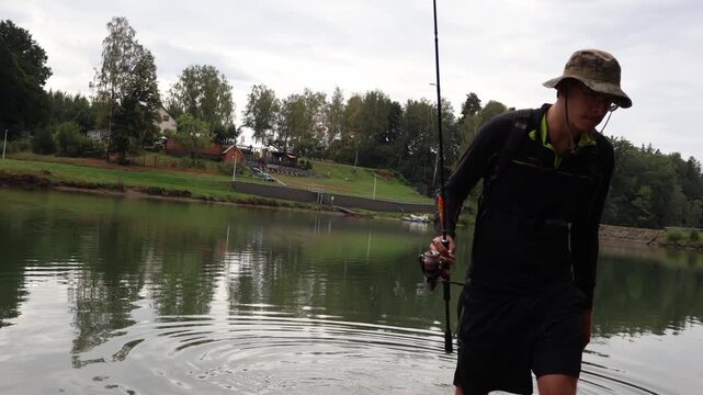 Angler wading through shallow lake water with fishing rod. Young fisherman targeting asp (Aspius aspius) during active outdoor sportfishing.