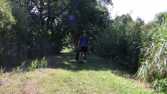 Fisherman walking towards camera carrying gear and chair near common reed (Phragmites australis). Angler arriving at nature fishing spot.