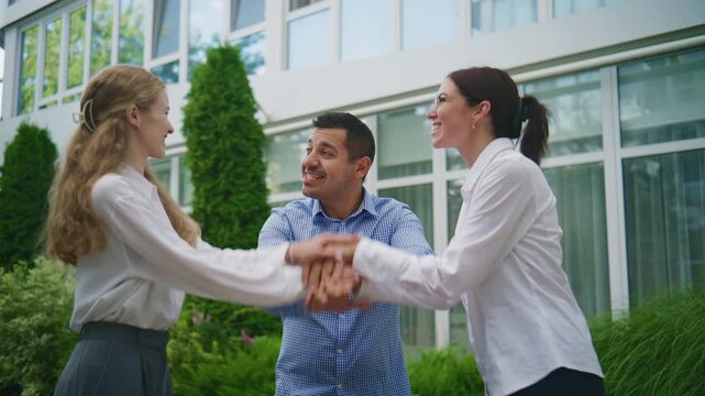 Three business colleagues stand outside a modern office building, stack their hands together, then raise them high with joyful energy, celebrating teamwork and unity