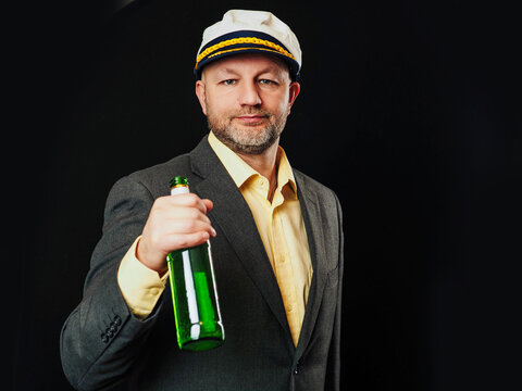 Man with grey beard wears captain's hat and suit. He stands confidently with green bottle in his hand. Costume party them. Black background. Funny male in his 40s or 50s. Enjoy drink concept.