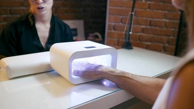 Professional nail technician providing a manicure service for a client. Client's hand drying gel nail polish in a modern UV lamp machine