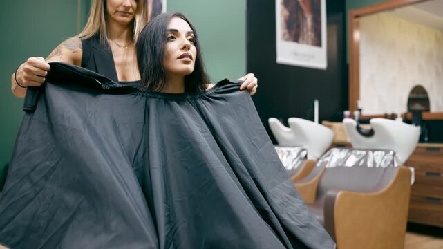 Professional hairdresser putting a black cape on a female client sitting in a chair. A woman preparing for a new hairstyle at the salon
