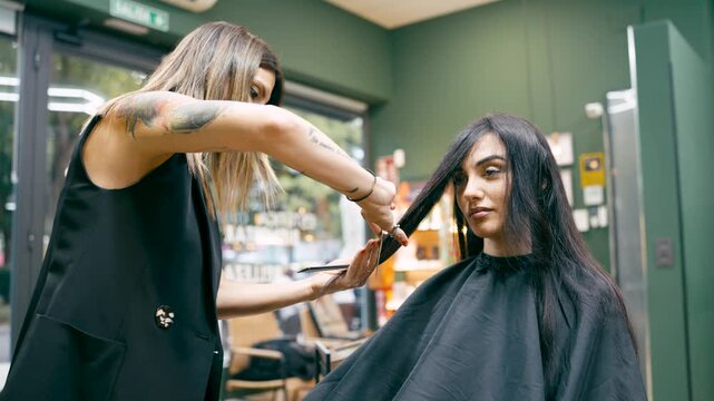 Female hairdresser with tattoos using scissors and comb to cut a young woman's long dark hair. Professional hair care and styling concept