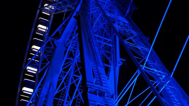Ferris wheel illuminated blue at night. Glowing blue ferris wheel structure stands tall against a dark night sky, its intricate metal framework illuminated, offering view of engineering and artistry