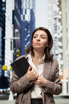Businesswoman carries a digital tablet while crossing city blocks toward an office building, walking along a metropolitan avenue with a digital touchscreen device during her work commute.