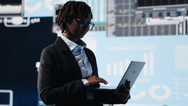 Manager uses laptop software to compare company metrics on a digital videowall before making decisions, analyzing key performance indicators.