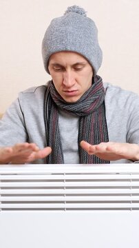 Chilled man wearing a hat and scarf sitting indoors, rubbing his frozen hands over a modern electric radiator to get warm during the central heating shutdown or a winter cold snap