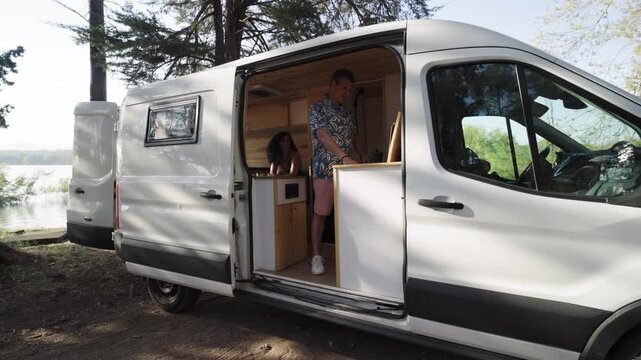 Mature couple cooking breakfast together inside their custom camper van. Nomadic lifestyle on a road trip near a lake during summer vacation