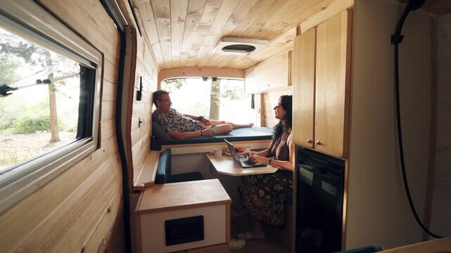 Mature couple relaxing inside their custom camper van. Woman working on laptop while man rests on bed using phone. Digital nomad lifestyle