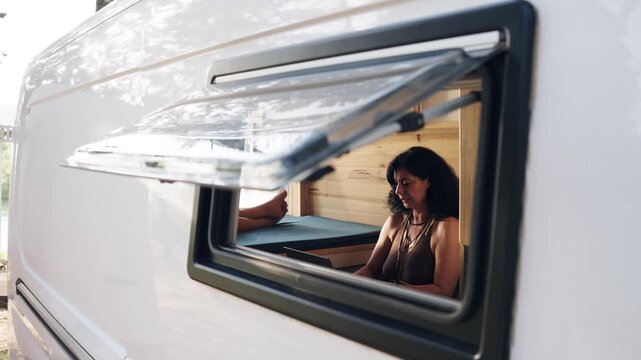 Outside view of a senior woman working on a laptop inside her camper van. Digital nomad enjoying the freedom of remote working on vacation