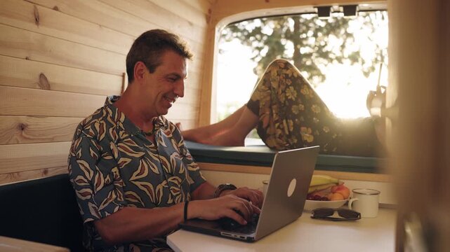 Happy mature man working on a laptop inside a camper van while his partner relaxes on the bed. Digital nomad couple enjoying freedom