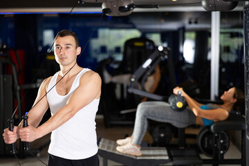 Fototapeta premium Sportive young man doing arm exercises on adjustable cable training device in well-equipped gym