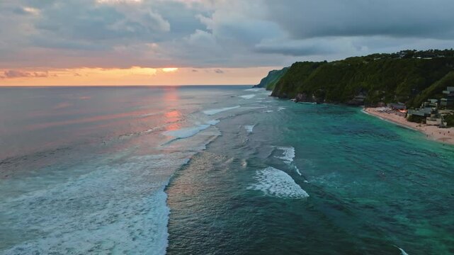 Aerial drone flight over tropical coastline of Bukit Peninsula, Uluwatu, Bali, during a colorful sunset, with rolling ocean waves meeting the sandy beach and lush green cliffs. Natural background