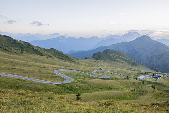 Scenic winding mountain road crossing green alpine slopes with layered mountain peaks and a wide valley in the distance.