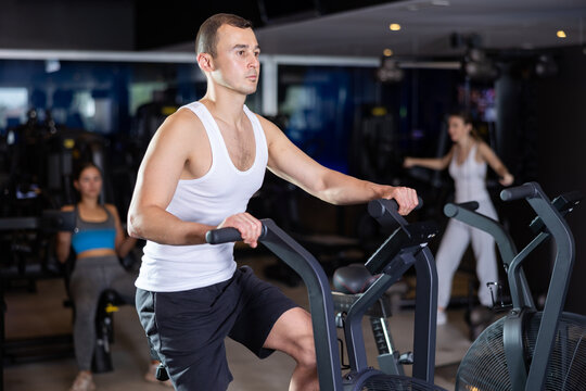 Determined young guy training with air bike in fitness hall during full-body workout