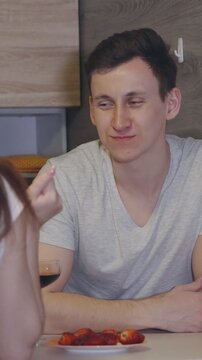 Young loving couple sharing a romantic moment at home, a caring girlfriend feeding her boyfriend a fresh strawberry and clinking glasses of red wine during a date night celebration