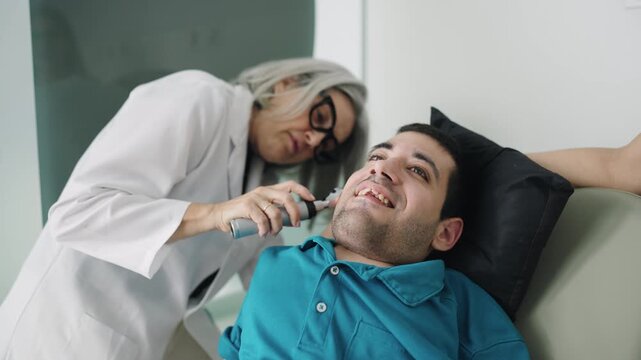 Female audiologist using a medical device to check the ear of a young adult male with spina bifida during a routine medical examination