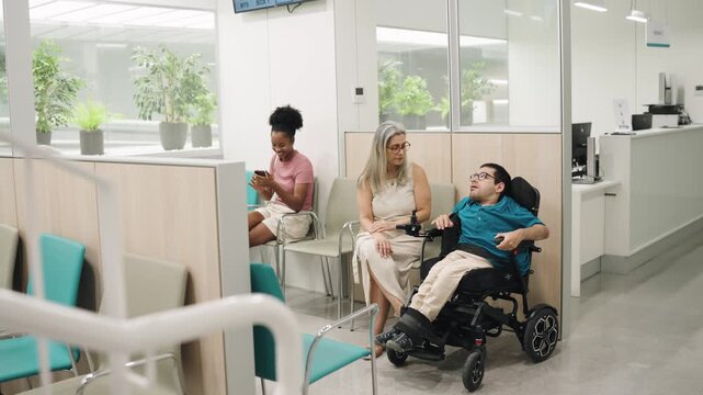 Young man with a physical disability sitting in an electric wheelchair, talking with his mother while waiting for a medical appointment