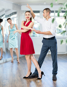Expressive young adult woman in elegant vibrant red dress dancing passionate bachata with male partner during dance class for adults led by female instructor..