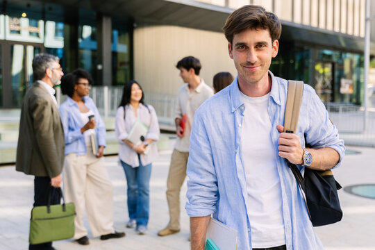 Portrait of a happy young caucasian university student carrying a bag outdoors, with a diverse group of classmates and professor blurred behind. Business school and education concept
